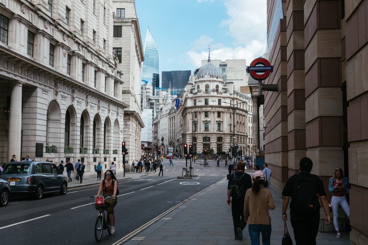 Una calle del centro de Londres: edificios de piedra clásicos a la izquierda, el rótulo circular del metro a la derecha, gente caminando por la acera, una ciclista en la calzada y un taxi negro aparcado, con una cúpula al fondo y una torre acristalada asomando entre los tejados.