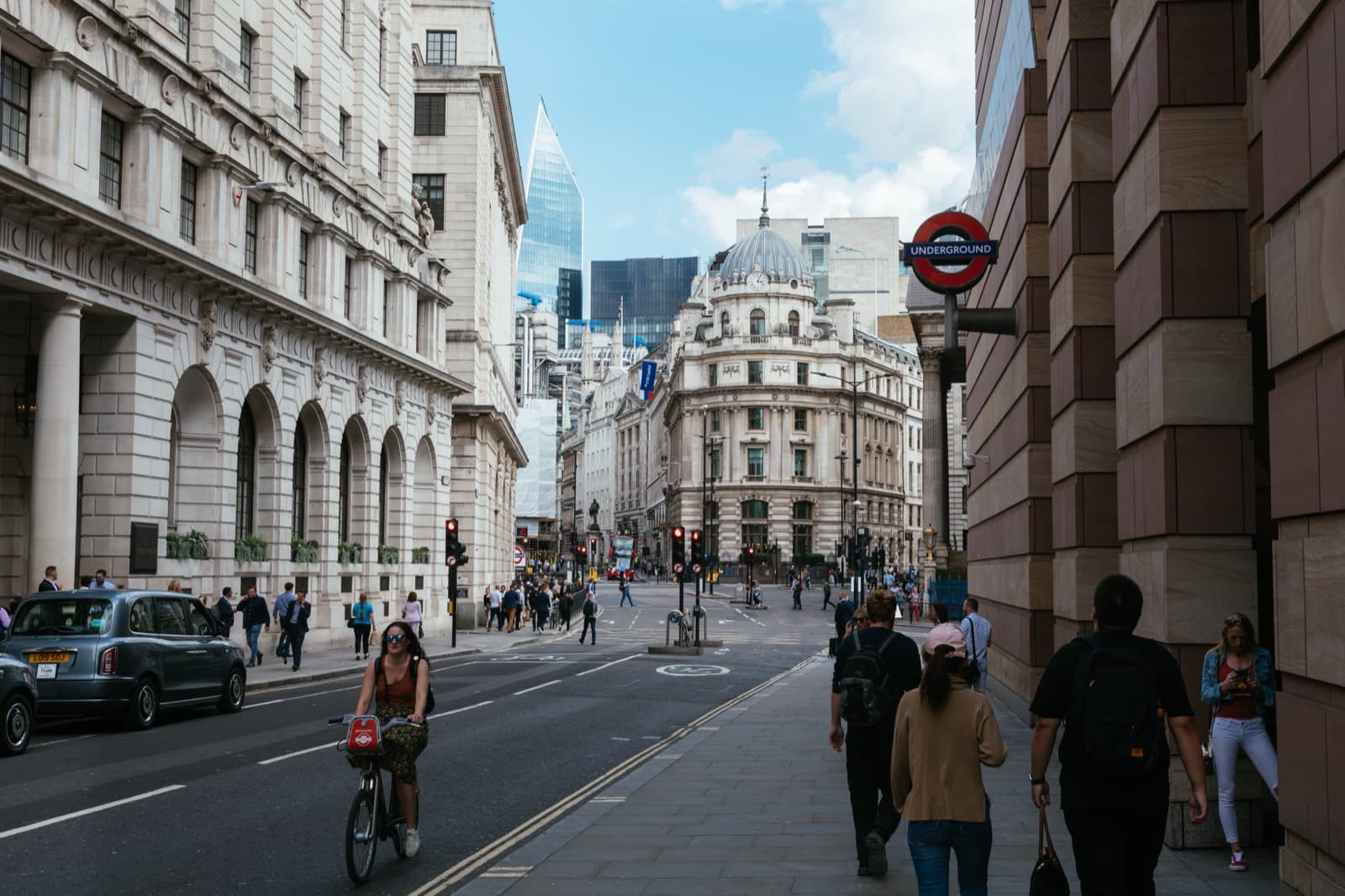 Una calle del centro de Londres: edificios de piedra clásicos a la izquierda, el rótulo circular del metro a la derecha, gente caminando por la acera, una ciclista en la calzada y un taxi negro aparcado, con una cúpula al fondo y una torre acristalada asomando entre los tejados.