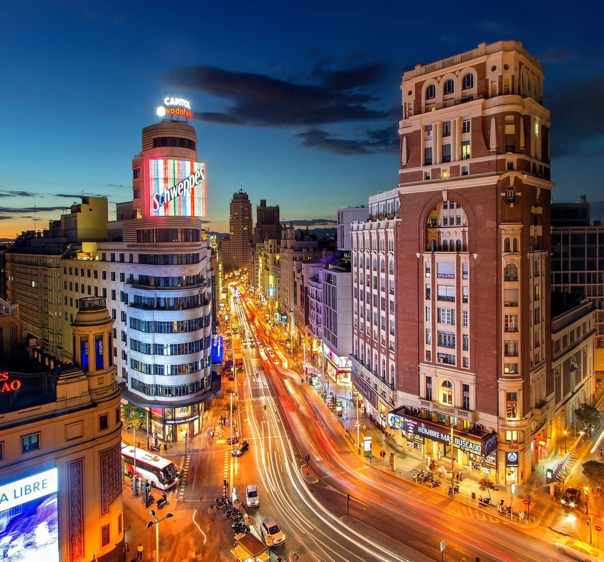 La Gran Vía de Madrid al anochecer, vista desde lo alto: el edificio Capitol con su rótulo luminoso, los comercios encendidos a ambos lados y los rastros de luz del tráfico bajando por la avenida bajo un cielo azul oscuro.