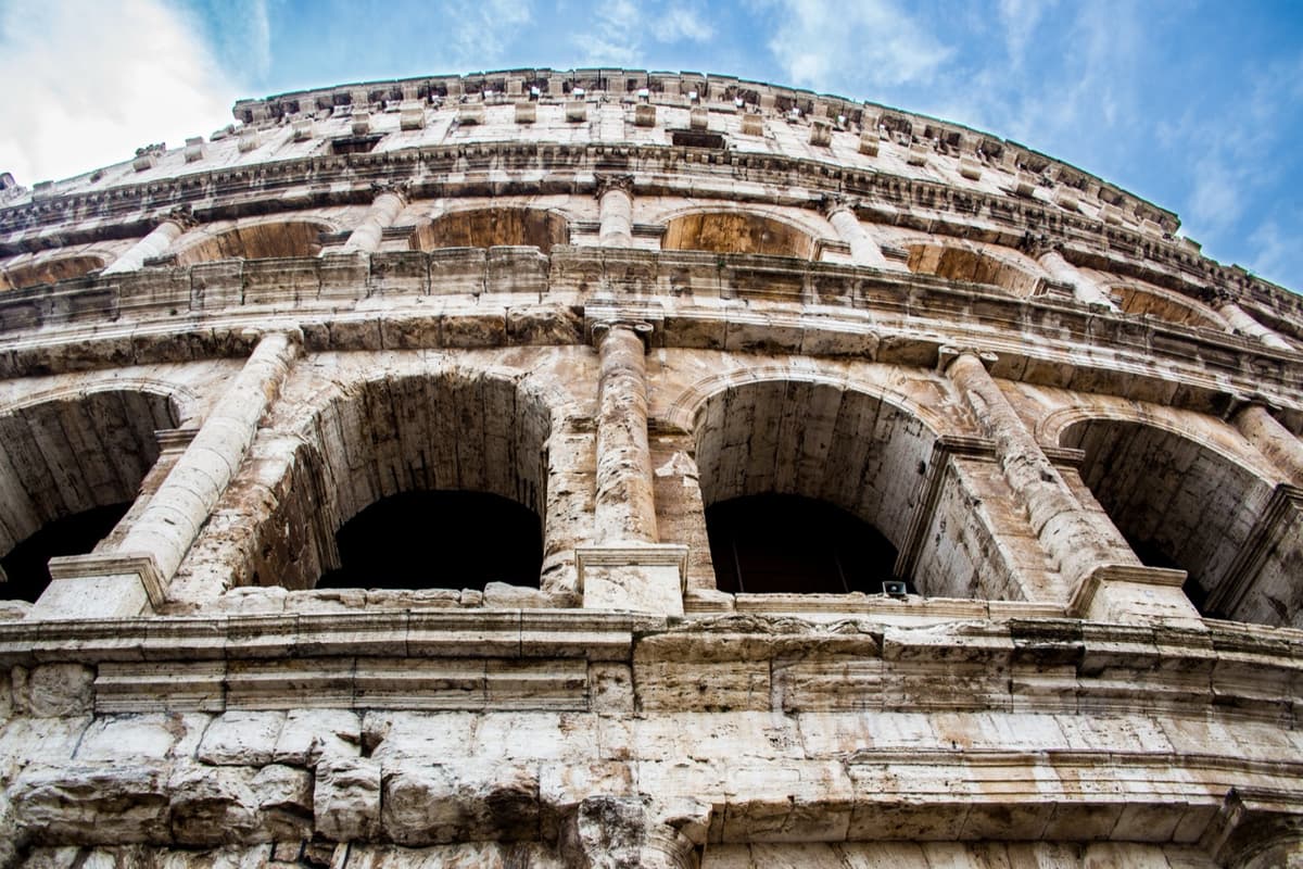 La fachada del Coliseo de Roma vista desde abajo en primer plano: la piedra clara y desgastada, dos arcos en sombra y las hileras de arquerías curvándose hacia arriba contra un cielo azul con nubes finas.