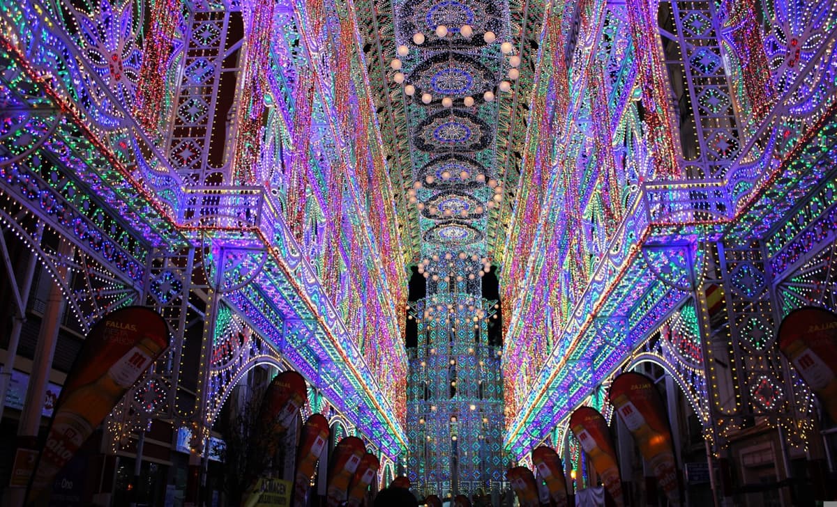 Una calle de Valencia cubierta de lado a lado por arcos de luces de colores durante las Fallas, con banderolas colgadas a ambos lados y la bóveda luminosa perdiéndose al fondo de la calle.