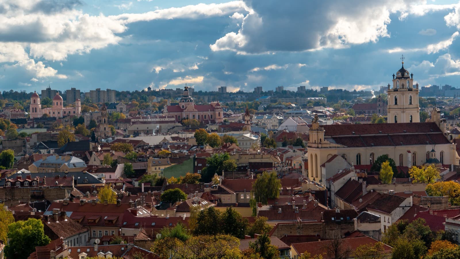Vista de la Ciudad Vieja de Vilnius desde lo alto: un mar de tejados de teja roja entre árboles con hoja amarilla de otoño, varios campanarios y torres de iglesia asomando entre las casas, y al fondo los bloques de la ciudad moderna bajo un cielo de nubes densas.